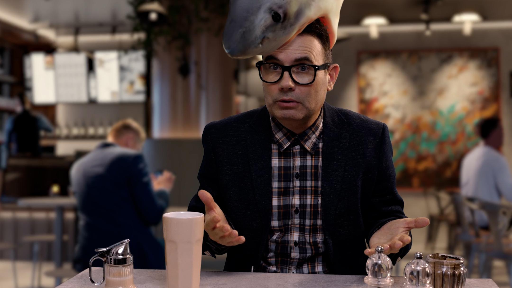 Eine Person sitzt an einem Tisch in einem modern eingerichteten Café oder Restaurant. Auf dem Kopf der Person befindet sich die realistisch dargestellte Figur eines großen Hais, der den Kopf von oben zu umschließen scheint. Vor der Person stehen ein helles Getränk in einem hohen Glas sowie mehrere Gewürzstreuer und eine kleine Karaffe. Im Hintergrund sind weitere Personen an Tischen zu sehen sowie eine Bar und dekorative Wandgestaltung.